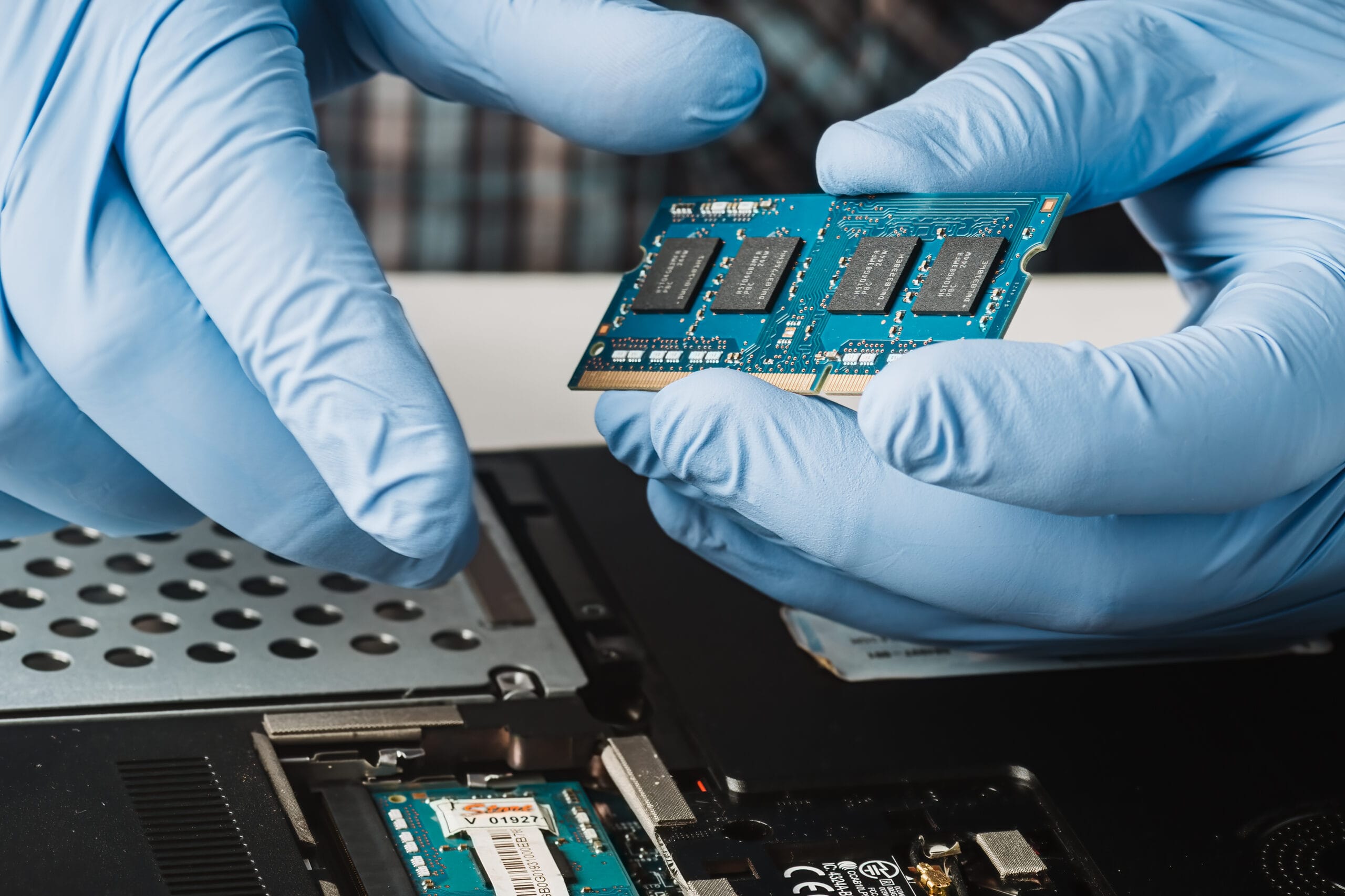 Hände mit Schutzhandschuhen halten ein RAM-Modul über einem geöffneten Computer